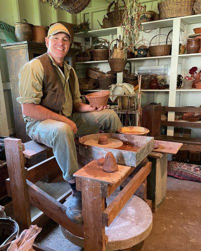 Kirke Martin smiling at his pottery wheel with some of his works on a shelf behind him